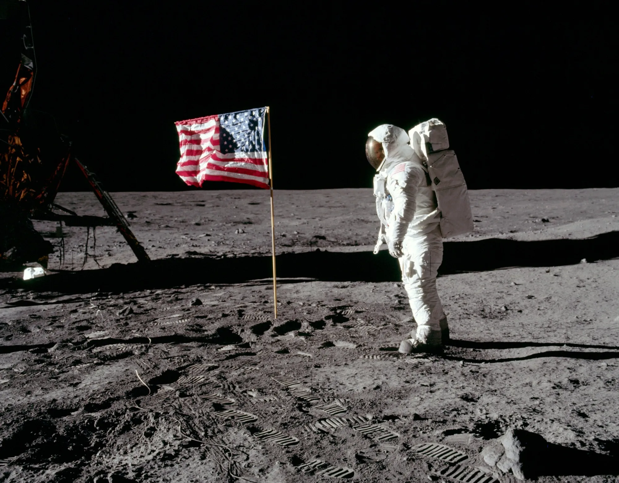 Fotografia oficial da missão Apollo 11 mostrando astronauta ao lado da bandeira dos EUA na superfície lunar. Análise de sombras e iluminação para o Portal Lumen Ancestral.